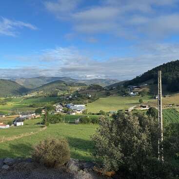 Uma paisagem rural tranquila com campos verdes, casas espalhadas, colinas onduladas, céu azul, nuvens fofas e vegetação exuberante. Linhas de energia e arbustos completam a vista panorâmica.