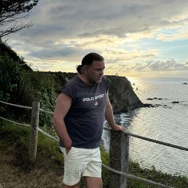 Um homem está próximo a uma cerca de corda em um penhasco com vista para o oceano ao pôr do sol, vestindo uma camisa "Polo Sport" e bermuda clara, com ar pensativo.