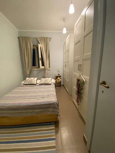 Um quarto aconchegante e estreito com uma cama de casal, um tapete listrado, duas almofadas, guarda-roupas brancos, um abajur e uma janela aberta com cortinas. Iluminação aconchegante.