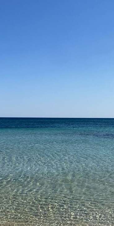 Águas rasas e cristalinas se estendem em direção a um mar calmo e de um azul profundo. O céu acima está sem nuvens, criando um cenário litorâneo tranquilo, sereno e revigorante.