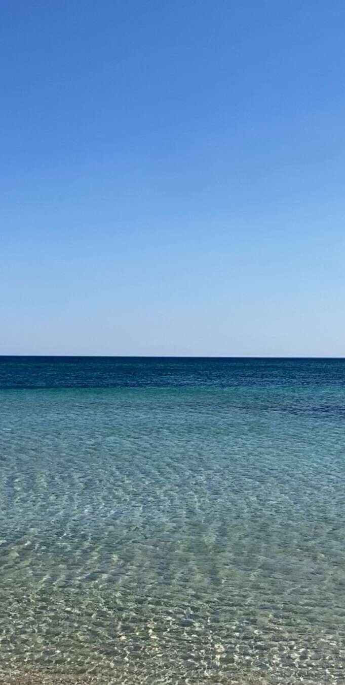 Águas rasas e cristalinas se estendem em direção a um mar calmo e de um azul profundo. O céu acima está sem nuvens, criando um cenário litorâneo tranquilo, sereno e revigorante.