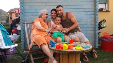Das Bild zeigt eine dreiköpfige Familie, bestehend aus einem Mann, einer Frau und einem Baby, die auf einer Holzbank vor einer blauen Wand sitzen, mit einem Tisch voller Spielzeug im Vordergrund.