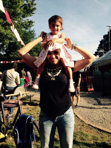 Das Bild zeigt eine Frau, die ein junges Mädchen auf den Schultern trägt. Im Hintergrund sind Bäume und ein blauer Himmel zu sehen.