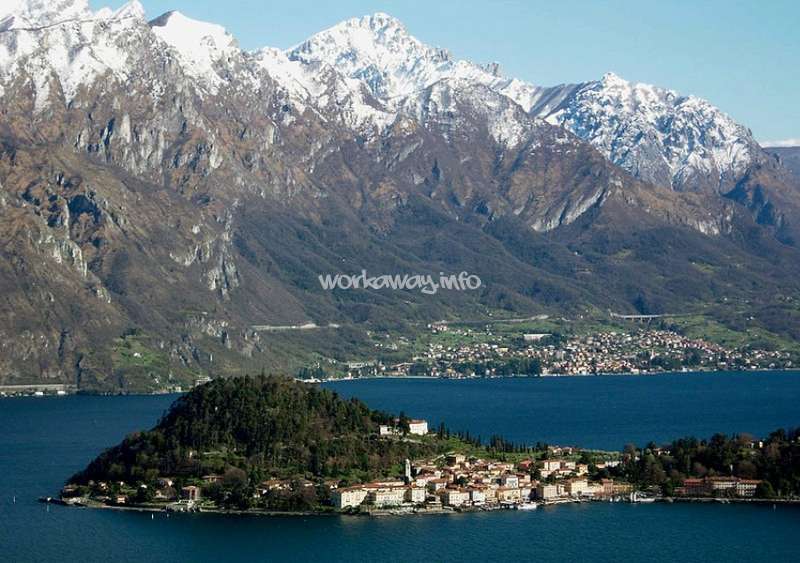 Das Bild zeigt eine ruhige Seeszene mit einer kleinen Stadt auf einer Insel, umgeben von Bergen und einem klaren blauen Himmel.