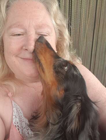 A woman with light curly hair smiles as a black and brown dachshund lovingly licks her nose. They share a close, affectionate moment together outdoors.