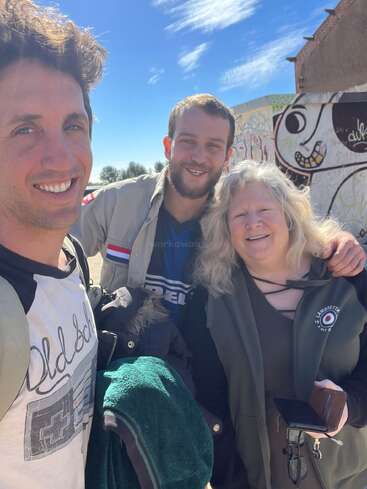 Three people smiling outdoors on a sunny day. Behind them is a colorful wall with graffiti art. They look happy, enjoying a casual and friendly moment together.
