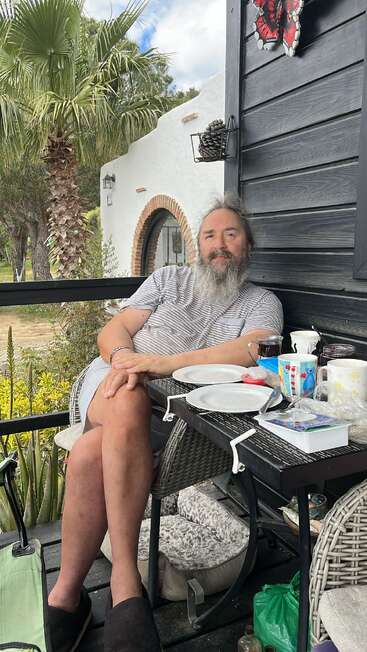 A bearded man relaxes on a patio, sitting comfortably by a table with dishes. Palm trees, plants, and a white building create a peaceful, outdoor atmosphere.