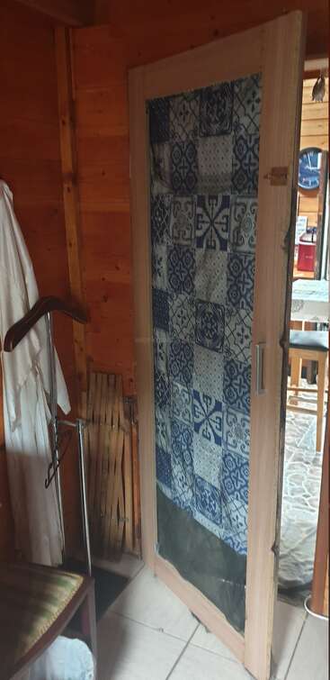 A wooden door with a blue, patterned fabric covering its glass panel stands open. Nearby are a coat hanger, folding chair, and rustic wooden walls.