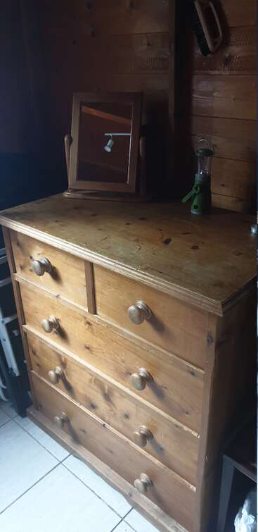 A wooden chest of drawers with round handles stands against a wood-paneled wall. On top, there's a mirror, green lantern, and a brush hanging nearby.