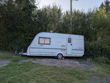 L'image représente un camping-car blanc garé sur un chemin de terre, entouré d'arbres et de buissons, avec une zone herbeuse au premier plan.