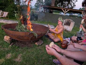 Un groupe est assis autour d'un grand feu de camp en plein air, jouant des instruments et portant des vêtements d'été. Une femme porte une couronne de fleurs, suggérant une réunion festive ou rituelle.