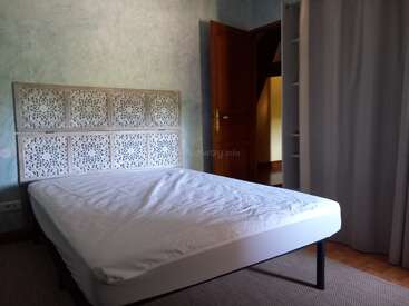 A simple bedroom with a carved decorative headboard, a bed with a white sheet, light green textured walls, an open wooden door, and partially visible closet.