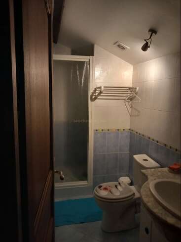 A dimly lit bathroom featuring a shower, toilet, sink, blue mat, towel rack with hangers, beige tiled walls, and minimal accessories on the counter.