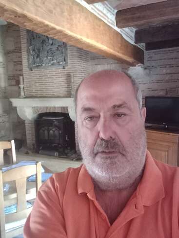 A man with a gray beard wearing an orange polo shirt takes a selfie inside a rustic room with wooden beams, stone fireplace, and wooden furniture.