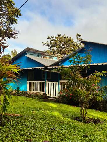 Ein leuchtend blaues Haus mit einer weißen Veranda steht inmitten von üppigem grünem Gras, tropischen Pflanzen und Bäumen unter einem strahlenden, teilweise bewölkten Himmel.