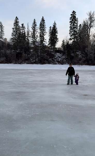 A person and a child stand together on a frozen, icy surface. Pine trees and snow-covered ground create a chilly winter backdrop beneath a pale blue sky.