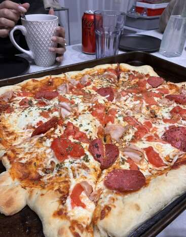 A large rectangular homemade pizza topped with cheese, pepperoni, tomatoes, and herbs sits on a tray. Nearby, someone stirs a drink beside a cola can.
