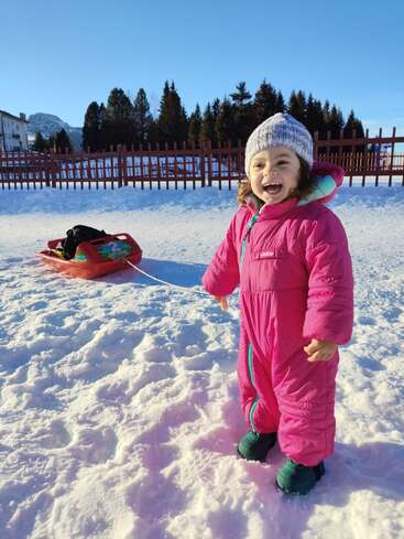 Uma criança feliz, vestindo um macacão rosa brilhante, está na neve, sorrindo muito, puxando um trenó vermelho. Árvores perenes e montanhas aparecem no fundo ensolarado.