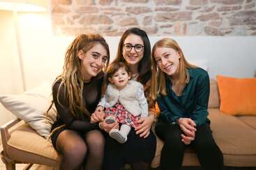Quatro mulheres, incluindo um bebê, sentam-se juntas em um sofá aconchegante, sorrindo calorosamente. O fundo apresenta uma parede de tijolos e iluminação suave, criando uma atmosfera acolhedora.