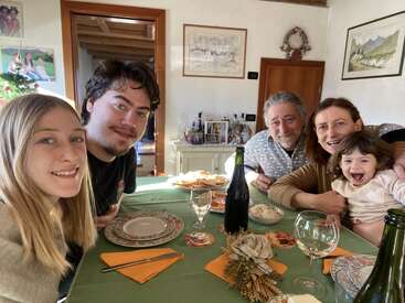 A family of five smiles at a dining table set for a meal, enjoying each other’s company in a bright, cozy home decorated with paintings and photos.