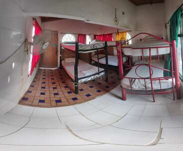 Dieses Bild zeigt ein kleines Zimmer mit zwei Stockbetten aus Metall, bunten Vorhängen, einem gefliesten Boden, einem Ventilator an der Wand und hellem Sonnenlicht, das hereinströmt.