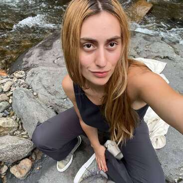 Una joven con el pelo largo y castaño claro se hace un selfie cerca de un arroyo rocoso. Lleva un top negro sin mangas, pantalones oscuros y zapatos informales al aire libre.