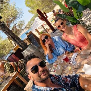 Trois amis portant des lunettes de soleil sont assis à la terrasse d'un restaurant en bord de mer ; ils profitent d'un verre et du soleil, entourés d'arbres et dans une ambiance estivale et détendue.