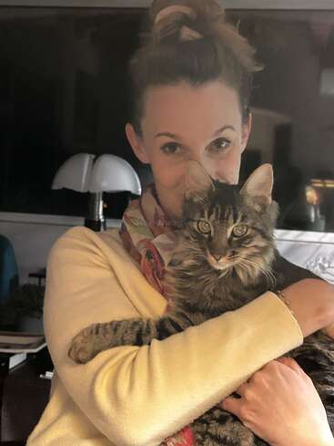 Une femme aux cheveux relevés en chignon, vêtue d'un pull de couleur claire, serre dans ses bras un chat tigré au pelage duveteux. Tous deux regardent l'objectif, créant ainsi un moment de tendresse.