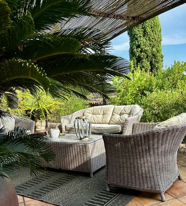 Un coin salon extérieur chaleureux, avec des meubles en osier sous une pergola ombragée, entouré de plantes et d'arbres luxuriants, créant une atmosphère relaxante et accueillante.