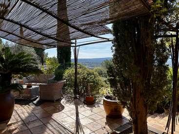 Une terrasse chaleureuse, agrémentée de chaises en osier, de plantes en pot et d'une pergola en bambou, donne sur une végétation luxuriante et des collines lointaines, sous un ciel bleu clair et ensoleillé.