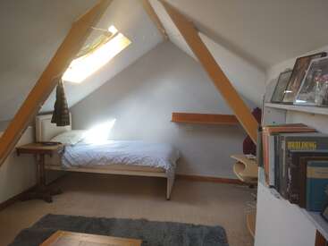 A imagem mostra um quarto aconchegante no sótão com uma cama de solteiro, vigas de madeira e uma pequena escrivaninha, com uma parede branca e uma janela com luz natural.