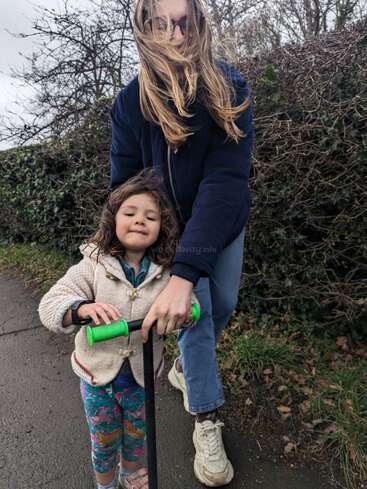 Uma mulher com longos cabelos loiros está ajudando uma menina com cabelos castanhos encaracolados a andar de scooter em um caminho pavimentado.