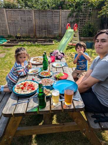 A imagem mostra uma família de três pessoas desfrutando de uma refeição em uma mesa de piquenique em um quintal, cercada por uma atmosfera serena e vegetação exuberante.