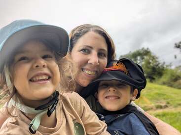 A imagem mostra uma mulher posando com duas crianças em um campo gramado, com as crianças usando chapéus e a mulher sorrindo para a câmera.