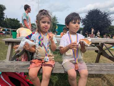 Duas crianças pequenas sentadas em um banco de piquenique, usando medalhas e saboreando lanches. Suas roupas estão desarrumadas, sugerindo atividades lúdicas ao ar livre em um evento ou festival de verão.