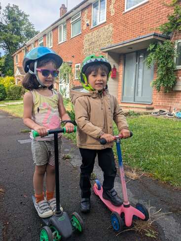 Duas crianças sorridentes, usando capacetes, estão em patinetes do lado de fora de uma casa de tijolos. Uma usa óculos escuros e shorts listrados, a outra usa capacete de dinossauro e jaqueta bege, pronta para se divertir.