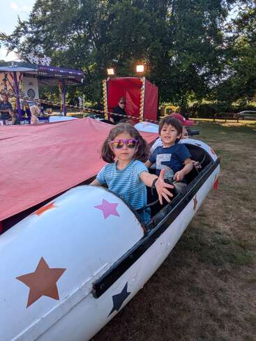 Duas crianças sentadas em uma atração de carnaval decorada com estrelas. A menina usa óculos escuros e estende a mão para frente, enquanto o menino sorri. Árvores, pessoas e barracas de feira os cercam.