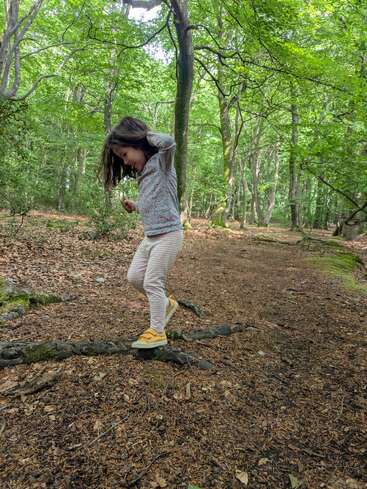 Uma menina se equilibra em uma raiz de árvore em uma floresta iluminada pelo sol. Ela está sorrindo, usando calças listradas e sapatos amarelos, cercada por vegetação e folhas caídas.