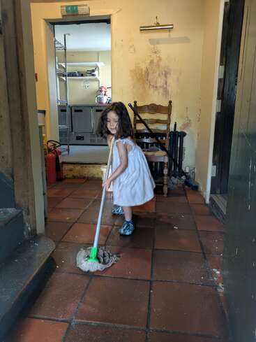 Uma jovem com vestido branco e sapatos azuis está limpando o chão de ladrilhos em uma sala um pouco bagunçada com paredes velhas e equipamentos de cozinha.