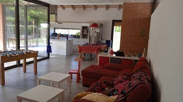 La imagen muestra un salón con una mesa y sillas, una gran ventana y una chimenea. Una estantería y una planta completan la estética de la habitación.