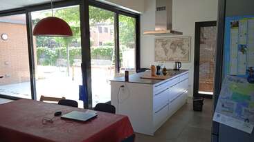 La imagen muestra una zona de cocina con mesa y sillas, con una gran ventana, un fregadero y un hornillo, con una mesa de comedor y sillas al fondo. La habitación está bien iluminada y organizada.