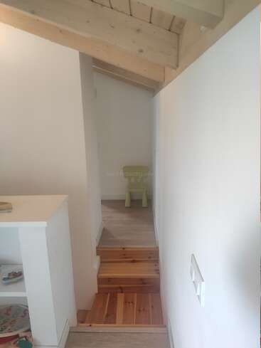 Ein kleiner, schmaler Flur mit hellen Holzböden und Stufen, weißen Wänden, freiliegenden Holzbalken an der Decke und einem grünen Stuhl am Ende neben einer Tür.