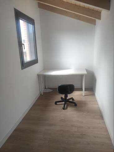 Dieses Bild zeigt einen kleinen, minimalistischen Raum mit weißen Wänden, Holzfußboden, einem einfachen Schreibtisch, einem Drehstuhl, einem Fenster und einer schrägen Holzdecke.