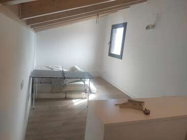 Ein minimalistisches Loft mit schräger Holzdecke, einem einzigen Fenster, einem einfachen Bett, einem klappbaren Tisch, weißen Wänden und einem kleinen Tresen mit Dekorationsgegenständen.