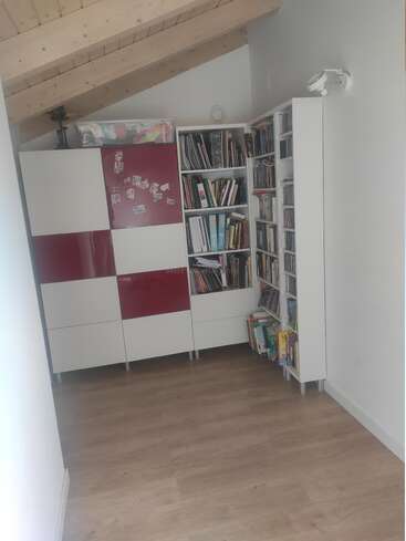 Dieser gemütliche Raum verfügt über schräge Holzbalken an der Decke, einen hellen Holzboden, moderne weiße und rote Schränke, Bücherregale, die mit Büchern, CDs und einigen Dekoartikeln gefüllt sind.