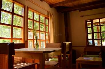 Sonnenlicht strömt durch große Holzfenster und taucht einen gemütlichen, rustikalen Raum in helles Licht. Ein schlichter Holztisch mit Stühlen und einer Vase mit Blumen lädt zu entspannter Ruhe im Haus ein.
