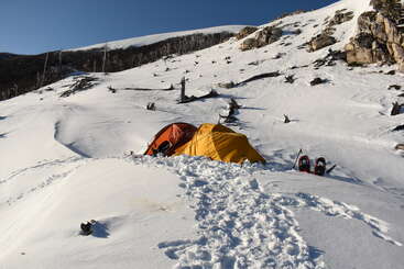 Ein leuchtend gelbes Zelt steht auf verschneitem Gelände, umgeben von Schneeschuhen und Bergausrüstung. Felsige Hügel und ein wolkenloser blauer Himmel runden diese friedliche Wintercamping-Szene ab.