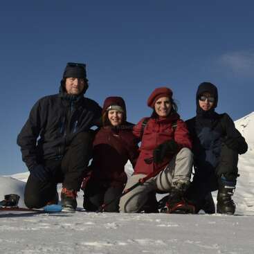 Cuatro personas, bien abrigadas para el invierno, están arrodilladas juntas sobre el suelo nevado. Detrás de ellas, el cielo azul y las colinas cubiertas de nieve crean un ambiente montañés fresco y alegre.
