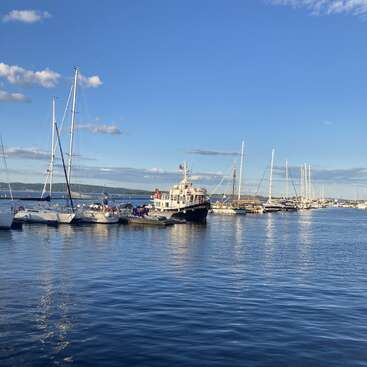 The image depicts a serene marina scene, featuring a large boat at the center, surrounded by smaller vessels, set against a backdrop of a blue sky with scattered clouds.