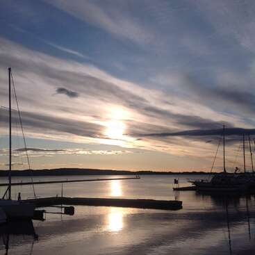 The image depicts a serene sunset scene at a marina, featuring boats docked in the water and a vibrant sky with clouds, reflecting off the calm surface.